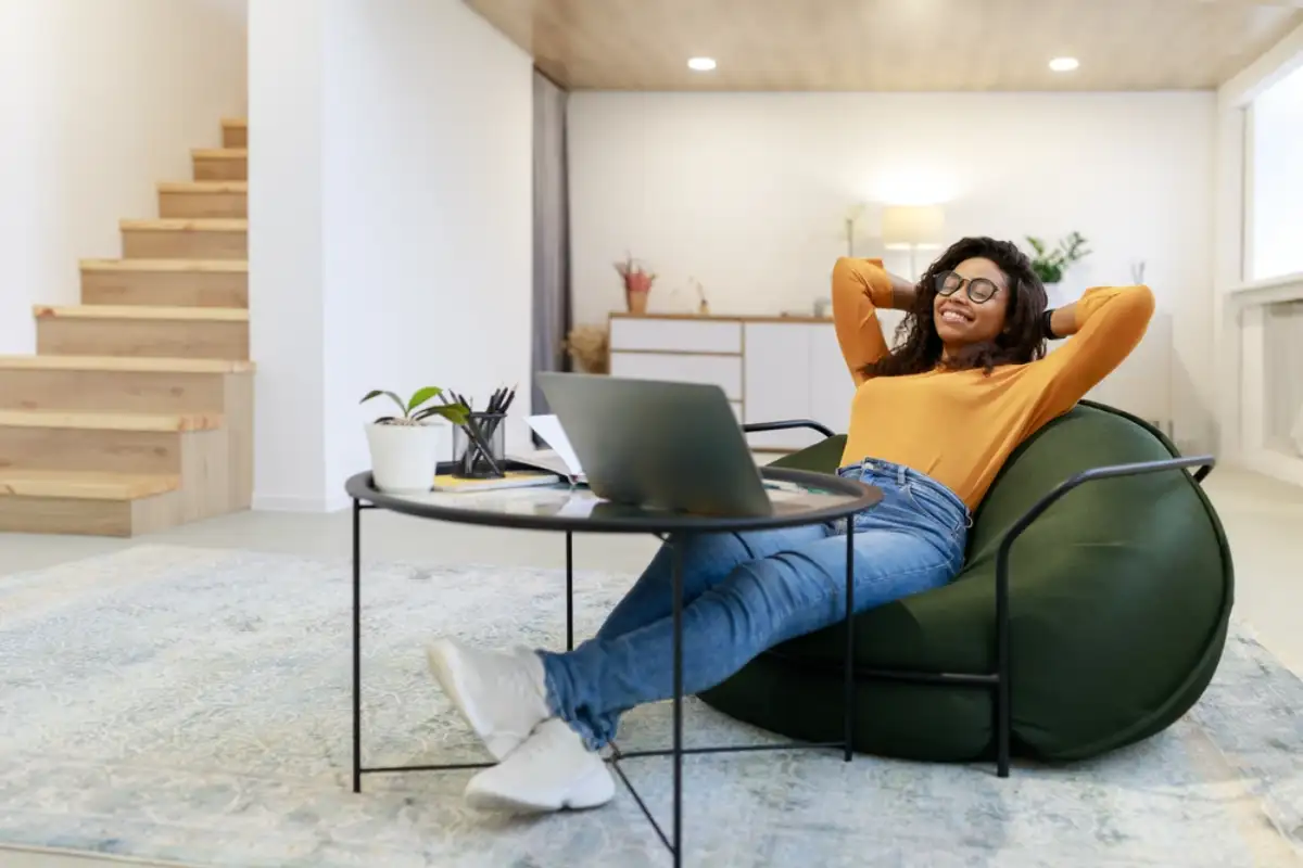 Smiling young black woman in glasses relaxing on bean bag chair sitting at tea table and resting, using pc laptop