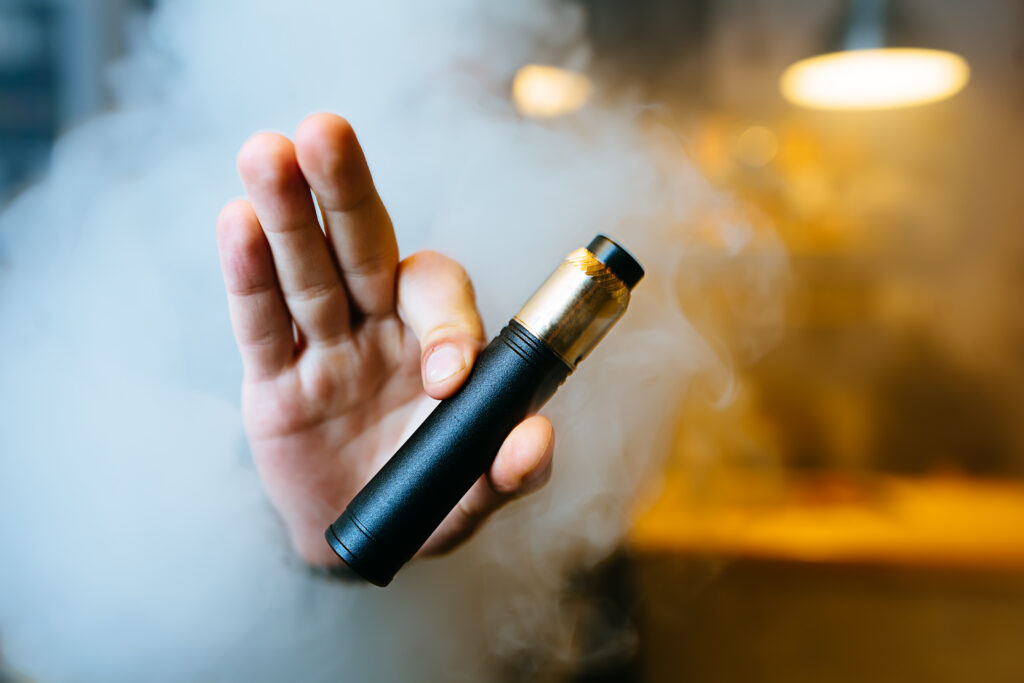 Young beard man show vaping device on his outstretched hand through a cloud of steam. Selective focus. Vaping concept. Copy space.